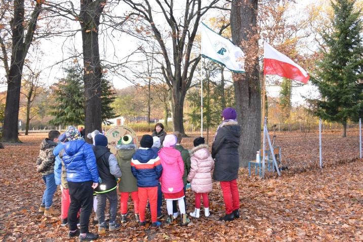 Obchody&#x20;Święta&#x20;Niepodległości&#x20;-&#x20;gra&#x20;terenowa&#x20;w&#x20;Szkole&#x20;Podstawowej&#x20;w&#x20;Brodach&#x2c;&#x20;fot&#x2e;&#x20;Paweł&#x20;Mrowiński&#x0a;Na&#x20;zdjęciu&#x20;jest&#x20;widoczna&#x20;grupa&#x20;dzieci&#x20;z&#x20;wychowawczynią&#x20;stająca&#x20;między&#x20;drzewami&#x20;przed&#x20;leśniczką&#x20;prowadzącą&#x20;konkurs&#x2e;&#x20;Nad&#x20;grupą&#x20;powiewają&#x20;flagi&#x3a;&#x20;flaga&#x20;państwowa&#x20;Polski&#x20;i&#x20;flaga&#x20;Lasów&#x20;Państwowych&#x2e;