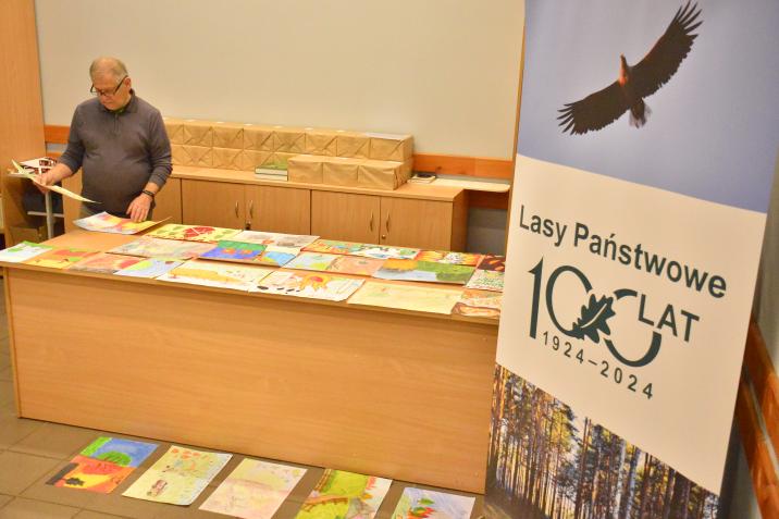 Sala&#x20;konferencyjna&#x20;OEPL&#x20;w&#x20;Jeziorach&#x20;Wysokich&#x20;z&#x20;drewnianym&#x20;blatem&#x20;prezentacyjnym&#x2c;&#x20;na&#x20;którym&#x20;rozłożone&#x20;są&#x20;kolorowe&#x20;prace&#x20;plastyczne&#x2e;&#x20;Po&#x20;lewej&#x20;stronie&#x20;stoi&#x20;artysta&#x20;plastyk&#x20;pan&#x20;Janusz&#x20;Orzepowski&#x2c;&#x20;który&#x20;ogląda&#x20;prace&#x2e;&#x20;Po&#x20;prawej&#x20;stronie&#x20;widoczny&#x20;jest&#x20;rollap&#x20;z&#x20;jubileuszowym&#x20;logo&#x20;100-lecia&#x20;Lasów&#x20;Państwowych&#x2e;&#x20;Na&#x20;górze&#x20;rollapu&#x20;znajduje&#x20;się&#x20;zdjęcie&#x20;lecącego&#x20;bielika&#x2e;&#x20;Fot&#x2e;&#x20;Paweł&#x20;Mrowiński